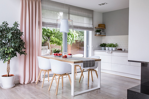 Cortinas romanas de gris y una cortina rosa en grande, ventanas de vidrio en una moderna cocina y comedor interior con una mesa de madera y sillas blancas photo
