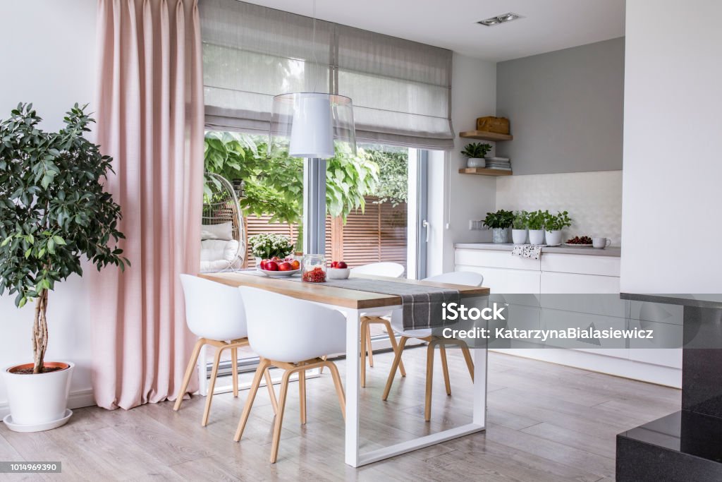 Gray roman shades and a pink curtain on big, glass windows in a modern kitchen and dining room interior with a wooden table and white chairs Curtain Stock Photo