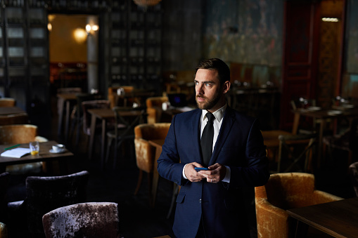 Serious pensive bearded restaurant owner in formalwear standing in empty establishment and texting message while looking away
