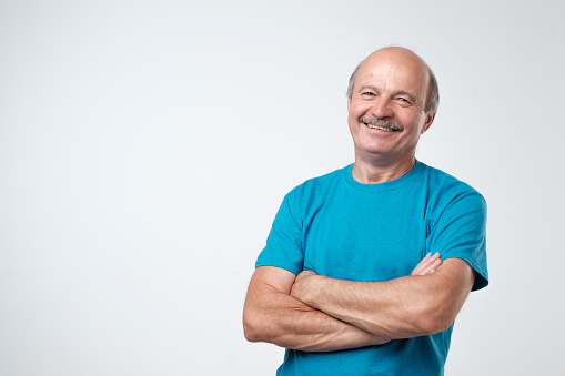 Charming handsome senior man in casual clothes keeping arms crossed and smiling while standing isolated on white background.