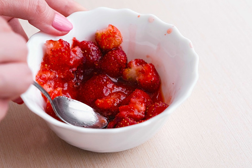 Woman kneads strawberries with a spoon and mixes it with sugar, making jam. Close-up hand