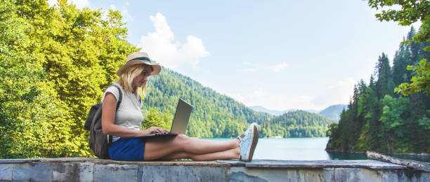 Beautiful girl traveler in a hat on a lake with a laptop Work outside the office stock photo