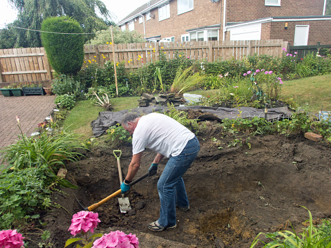 A senior man reconstructing a pond from a figure of eight to a rectangular shape using pick axe and spade.