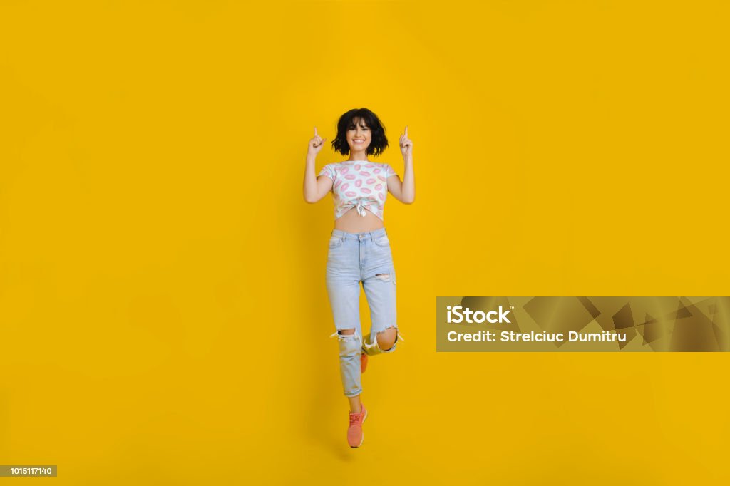 Cheerful woman jumping and pointing up Lovely young female in casual outfit smiling and pointing up while jumping on bright yellow background Women Stock Photo
