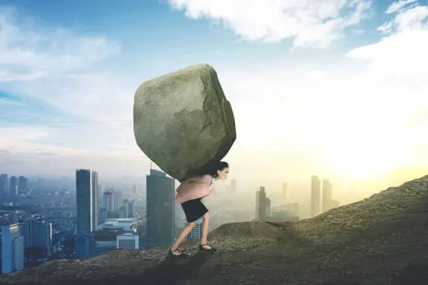 Picture of young Caucasian businesswoman lifting a big stone while climbing on the hill