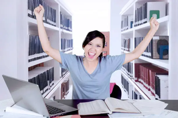 Picture of female college student looks excited while raising hands and studying in the library
