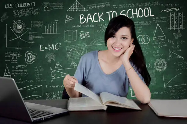 Picture of female college student smiling at the camera while reading a book and sitting with scribble in the chalkboard