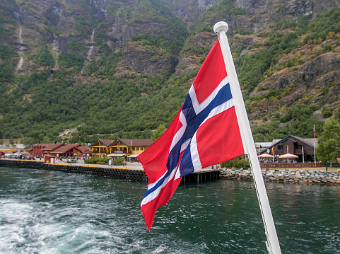 Norwegian flag is flying at the stern of a boat travelling through a fjord.