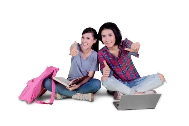 Two college students showing thumbs up while sitting together in the studio, isolated on white background