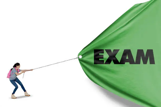 Picture of a beautiful girl struggling to pull a green banner with exam word, isolated on white background