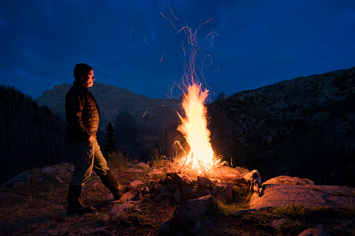 Adult male in down jacket and hat watches a fire to keep it under control on a cold night while camping at Elk Creek in the San Juan Mountains, Weminuche Wilderness, Rocky Mountains, Silverton, Colorado, USA