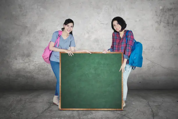 Two female college students smiling at the camera while holding a chalkboard with copy space