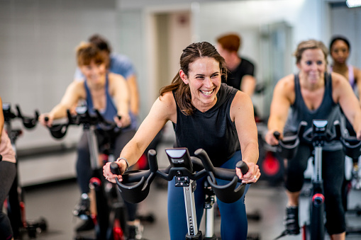 A group of adults are indoors in a fitness center. They are riding exercise bikes. One woman is smiling in the foreground.