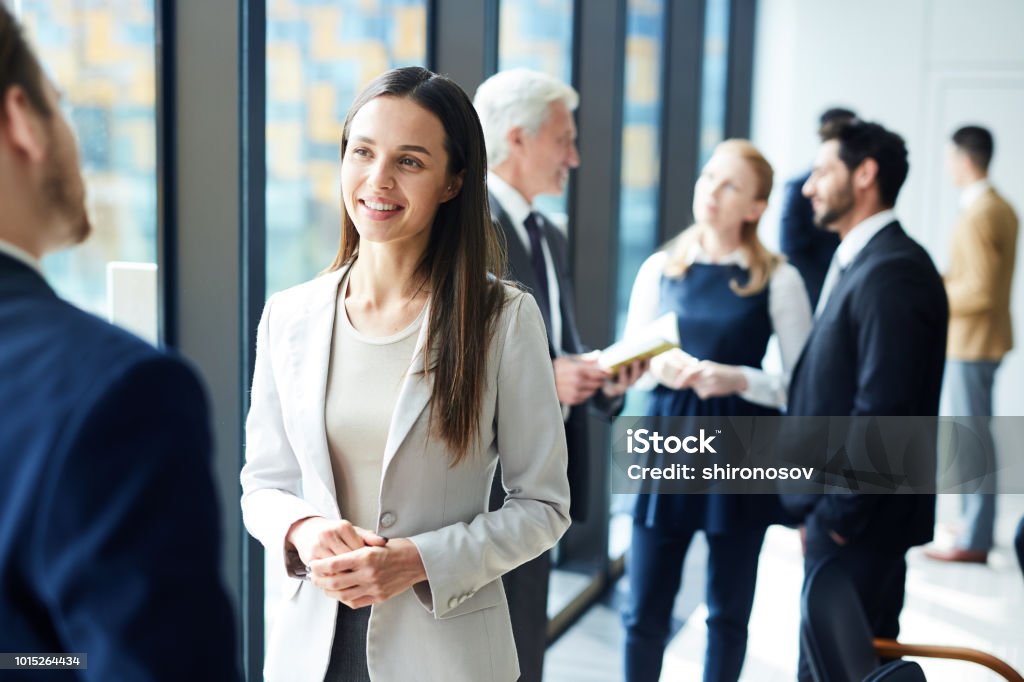 Pretty business lady talking to colleague Smiling pretty young business lady in jacket talking to colleague and discussing business forum topics during break Business Stock Photo