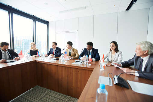 Cheerful confident multiethnic consuls in formalwear discussing future plans while participating in international debate at meeting