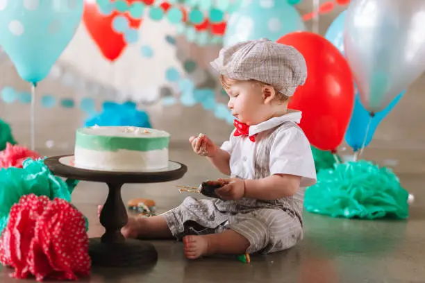 first birthday cakesmash. Adorable baby boy sitting on concrete floor and eating cake in decorated studio location.