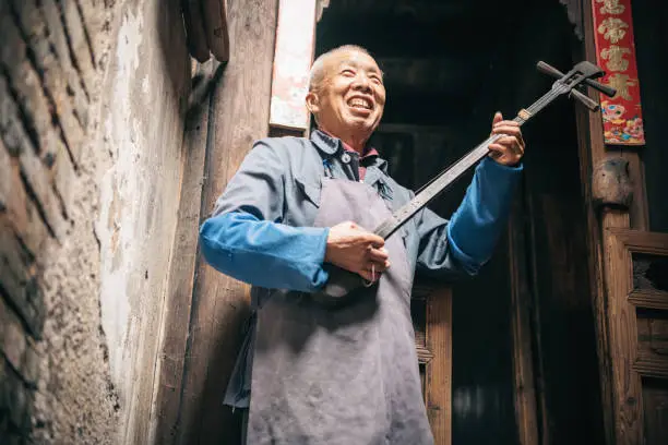 smiling chinese senior man playing longneck lute in front of wooden house in daxu ancient town