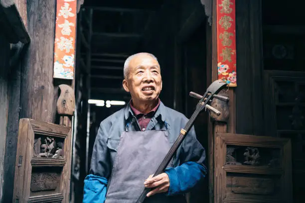 smiling chinese senior man holding longneck lute in front of wooden house in daxu ancient town