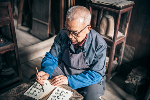 chinese senior manwriting chinese calligraphy characters with india ink on paper at little desk