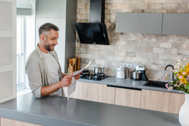 istock side view of smiling bearded man reading newspaper while standing at counter in kitchen 1015287634