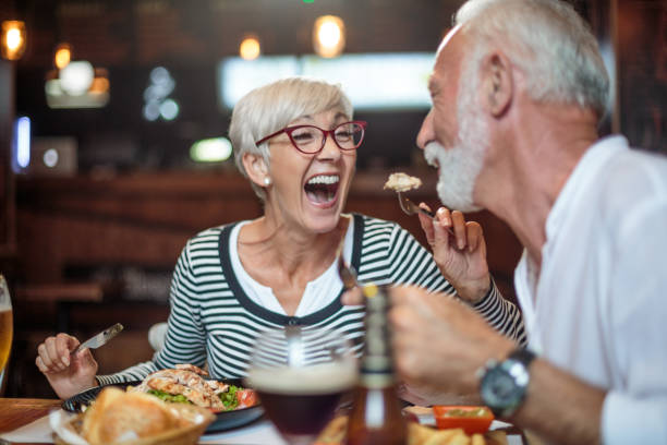 Senior woman laughing while feeding her male partner in the restaurant Active senior couple having a lunch in the restaurant and woman is feeding her partner. Drinks and food are on the table. barbecue social gathering photos stock pictures, royalty-free photos & images