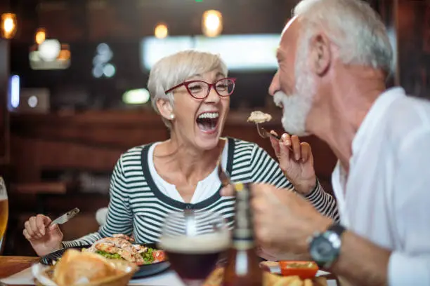 Active senior couple having a lunch in the restaurant and woman is feeding her partner. Drinks and food are on the table.