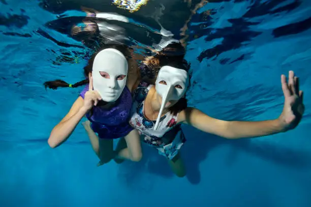 Underwater theater. Two unusual girls swim and play underwater in the pool on a blue background in white masquerade masks in beautiful dresses and look at the camera. Horizontal orientation.