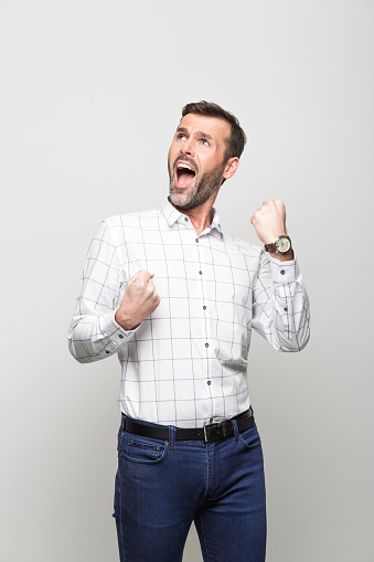 Portrait of handsome successful businessman wearing white shirt and jeans, shouting with raised fists. Studio portrait, grey background.