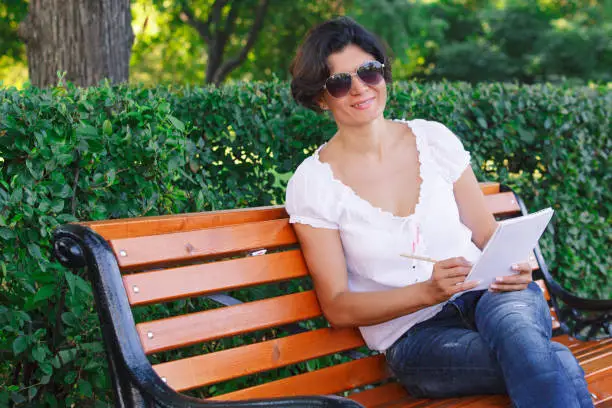 The girl smiles and writes in a notebook on a park bench. A middle-aged brunette woman in blue jeans and a white blouse.