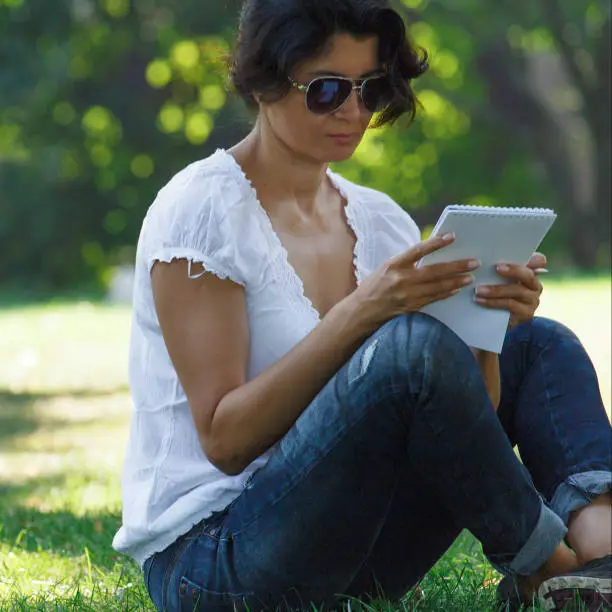 The brunette girl in the park is reading a record. A concept of calm mood and relaxation in a park on the green grass. A middle-aged woman in blue jeans.