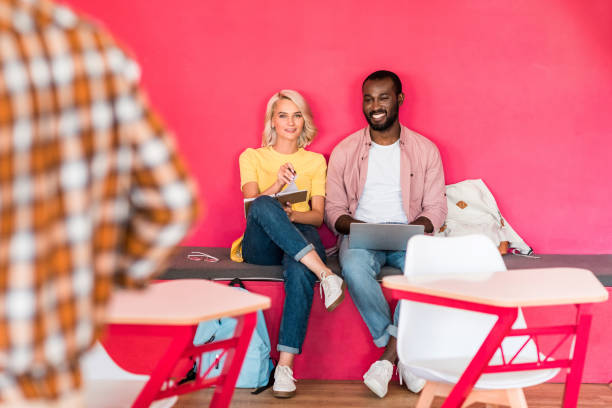 happy young students working with laptop while sitting under red wall