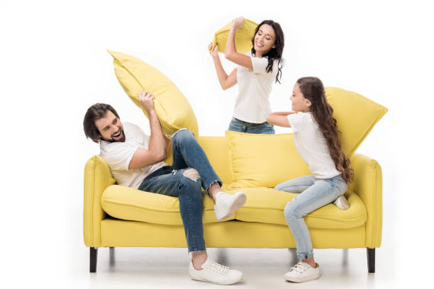 familia feliz en camisas blancas en amarillo sofá con almohadas aislado en blanco - lucha con almohada fotografías e imágenes de stock