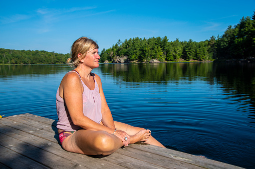 A pretty, mature woman sits on a dock by a beautiful lake looking out over the water with a slight look of anxiety or pensiveness.