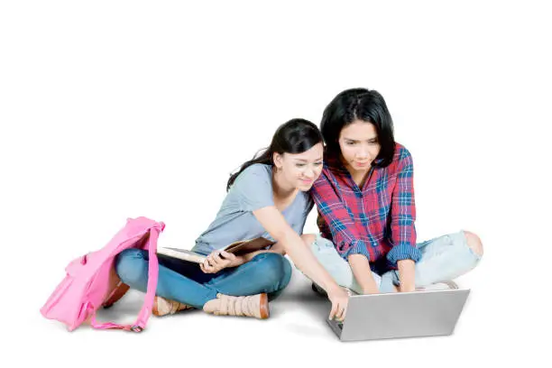 Picture of two female college students using a laptop while doing homework in the studio, isolated on white background