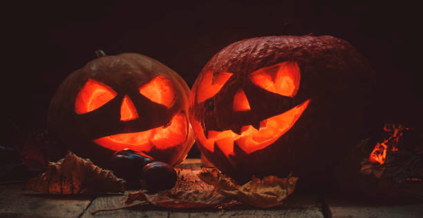 Two Halloween pumpkin Jack-o-Lantern on dark wooden background with fallen leaves and flames, selective focus and toned image Two Halloween pumpkin Jack-o-Lantern on dark wooden background with fallen leaves and flames, selective focus and toned image halloween pumpkin human face candlelight stock pictures, royalty-free photos & images