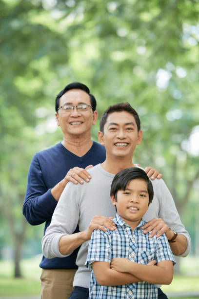 Confident Asian boy with father and grandfather in park Cute Asian boy smiling and keeping arms crossed while standing near cheerful father and grandfather on blurred background of park vietnamese old man portrait stock pictures, royalty-free photos & images