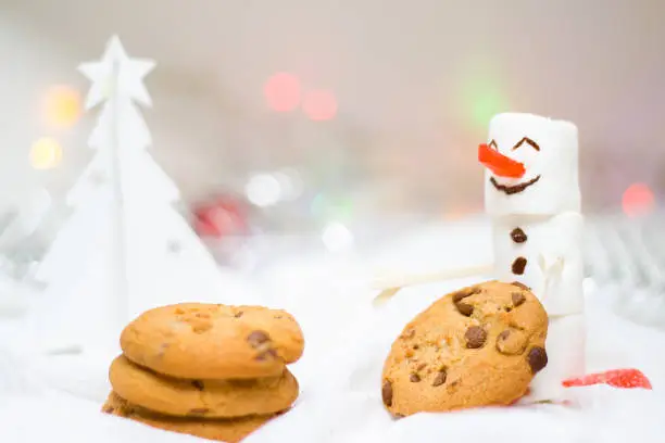 holidays, winter and celebration concept - Christmas and new year card with snowman from marshmallow, chocolate gingerbread cookies and white Christmas tree made of paper, blurred lights on background
