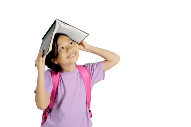 Image of pensive little girl putting a book on her head while standing in the studio, isolated on white background