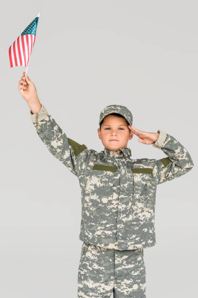 retrato de niño en camuflaje ropa saludando mientras sostiene el asta de la bandera americana en la mano aislado en gris - child patriotism saluting flag fotografías e imágenes de stock