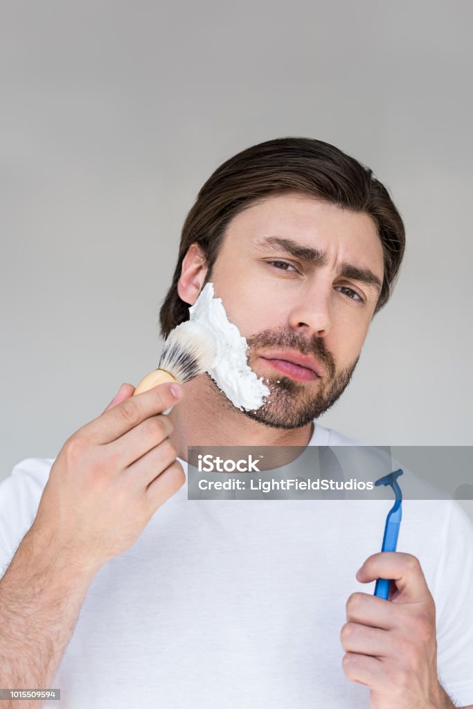 portrait of man with brush and razor in hands putting shaving foam on face on grey backdrop Adult Stock Photo