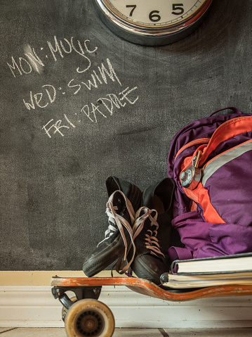 Everyday teenage belongings on the floor of the home interior. Long board, back pack and sport shoes against the wall with black board finish.