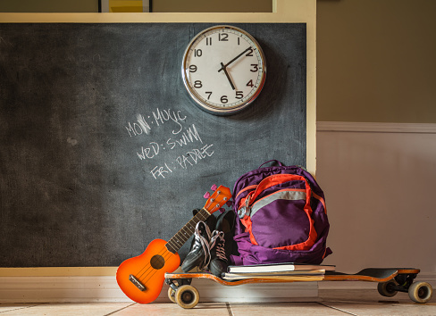 Everyday teenage belongings on the floor of the home interior. Ukulele, long board, , back pack and sport shoes against the wall with black board finish.