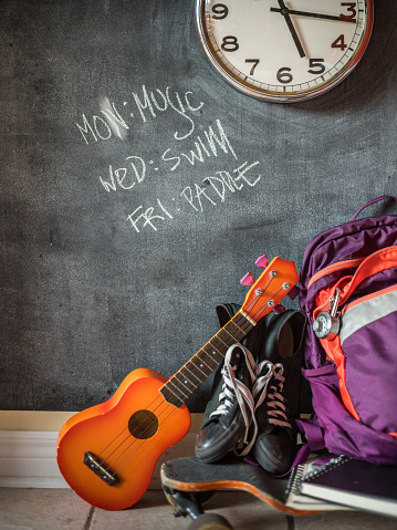 Everyday teenage belongings on the floor of the home interior. Ukulele, long board, , back pack and sport shoes against the wall with black board finish.