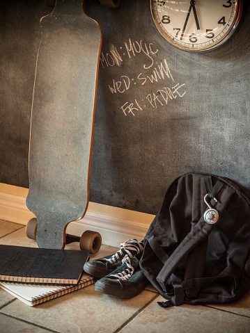 Everyday teenage belongings on the floor of the home interior. Long board, back pack and sport shoes against the wall with black board finish.