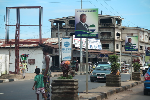 Political post-election Poster in Freetown, the capital of Freetowb on main street celebrating President Julius Bio having won the election in March  2018. There is a woman and a young child in front looking away from the camera, ready to cross the road as a car approaches. On the other side of the road a there is a woman in the distance with a large container worn on her head.   The picture was taken in late July 2018. There are people standing in the background, revealing a typical  West African urban atmosphere.