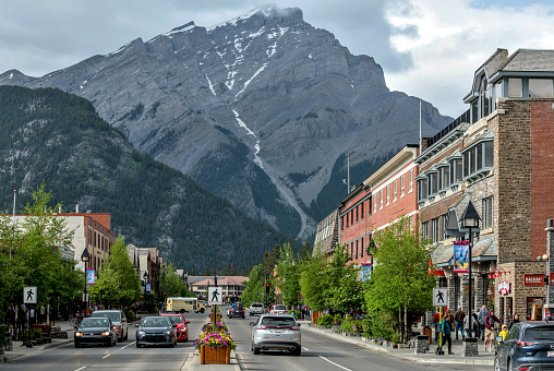 Banff, Alberta, Canada - June 9, 2018: Formidable Cascade Mountain towering high at far end of busy Banff Avenue on a cloudy Spring day in Downtown Banff. Banff National Park.