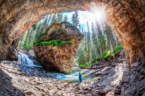 Hiker stands in awe of waterfall and limestone bedrock at a hidden cave in Johnston Canyon at Banff National Park, with sun bursting through the lush forest in the Canadian Rockies.