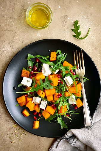 Delicious autumn pumpkin salad with arugula, feta cheese and pomegranate seeds on a black plate over beige slate, stone or concrete background.Top view.
