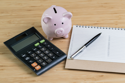 Pen with list of priority number on white note pad with pink piggy bank and calculator on wooden table as finance, money budget, saving or investment planning concept.