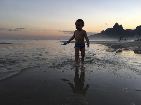 Toddler runs at the beach on a wet area while his reflection appears in the water. Ipanema Beach, Rio de Janeiro, Brazil. Boy wearing a speedo at low tide